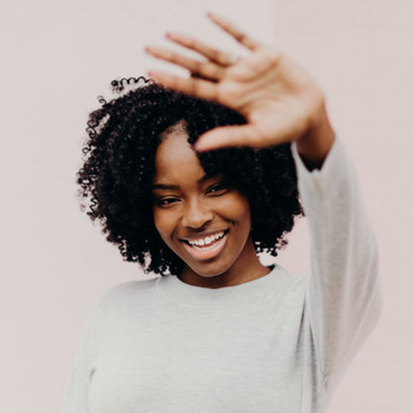 A happy young black woman in a casual grey sweater and jeans holds her hand up to camera while laughing and standing in front of a pink wall.