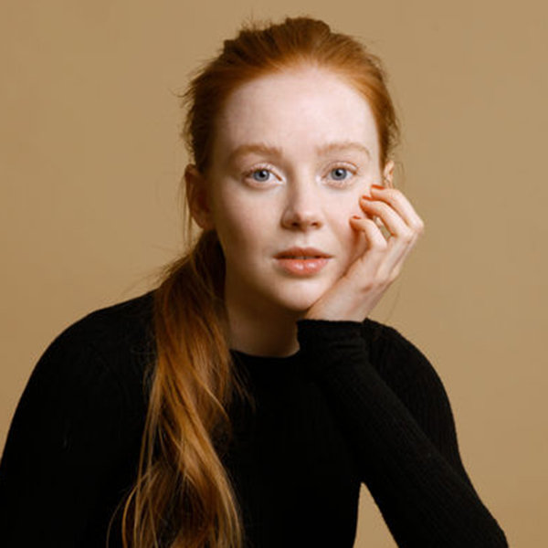 studio portrait of a young woman with long red hair, she's wearing a black sweater and black trousers, she leans on her hand and looks at the camera